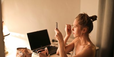 A young woman applies makeup at her vanity table, illuminated by soft light, in a cozy home setting.